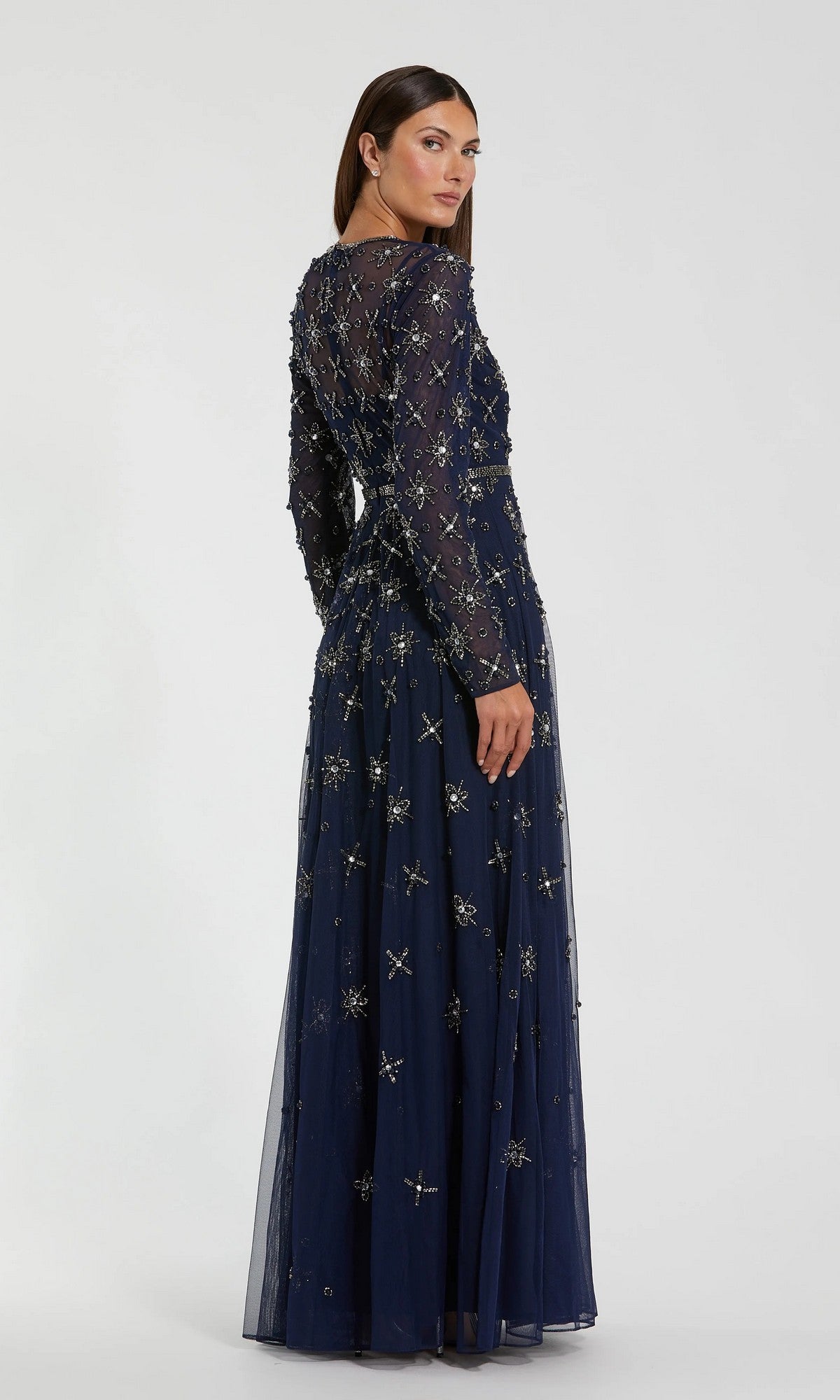 A woman stands facing away against a plain background, wearing the Mac Duggal 94216 long formal dress—a navy gown with long sleeves and intricate beaded embellishments.